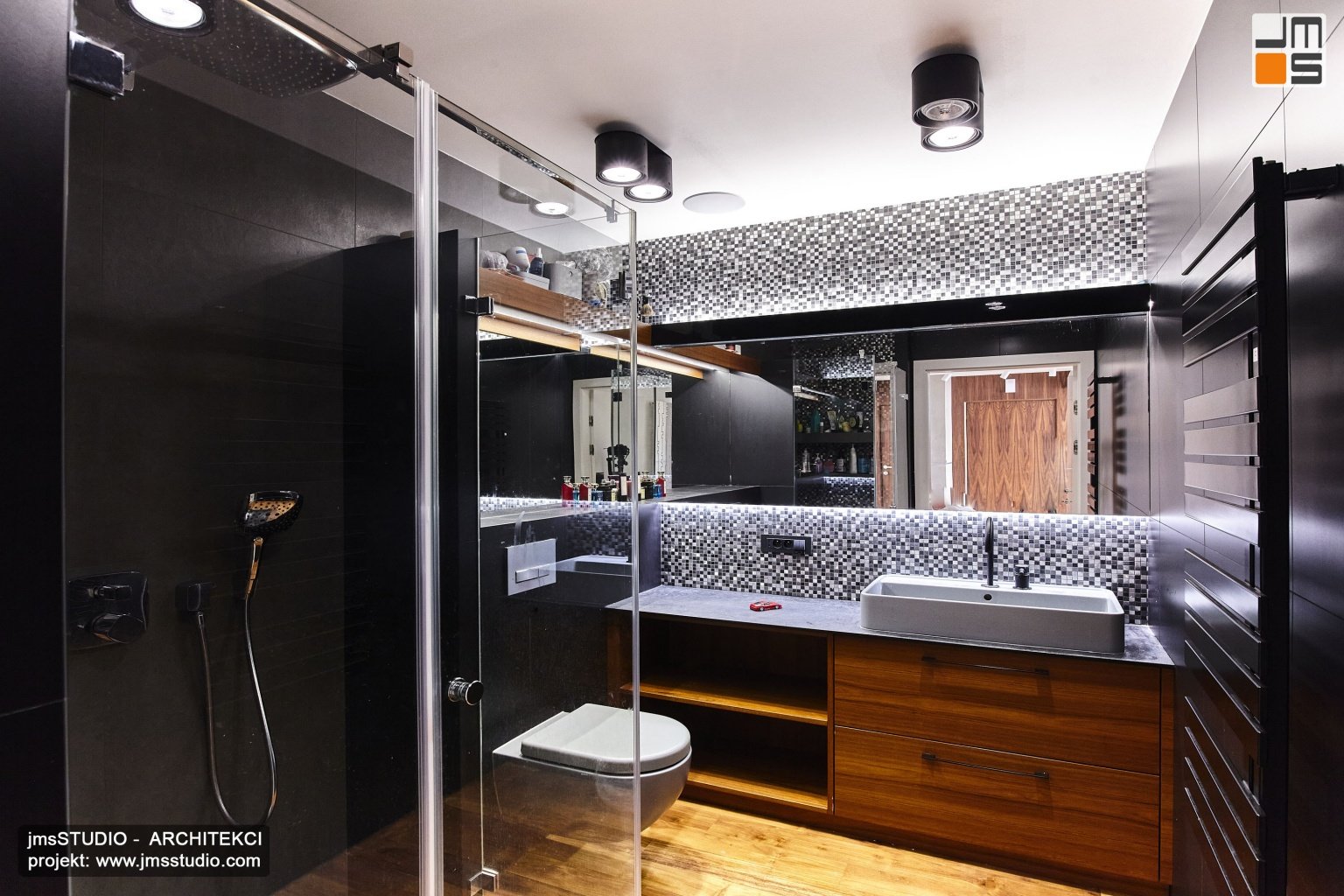 Projekt wnętrz pięknej ciemnej łazienki w apartamencie to pomysł na połączenie stalowej mozaiki z czarnymi płytkami i drewnem oraz technicznymi czarnymi lampami